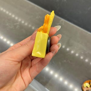 Puede incluir: Un peque&ntilde;o im&aacute;n tridimensional de color amarillo con forma de bolsa de la compra. La bolsa contiene elementos de color naranja que sobresalen por la parte superior. Un disco magn&eacute;tico negro circular est&aacute; fijado a la parte trasera.