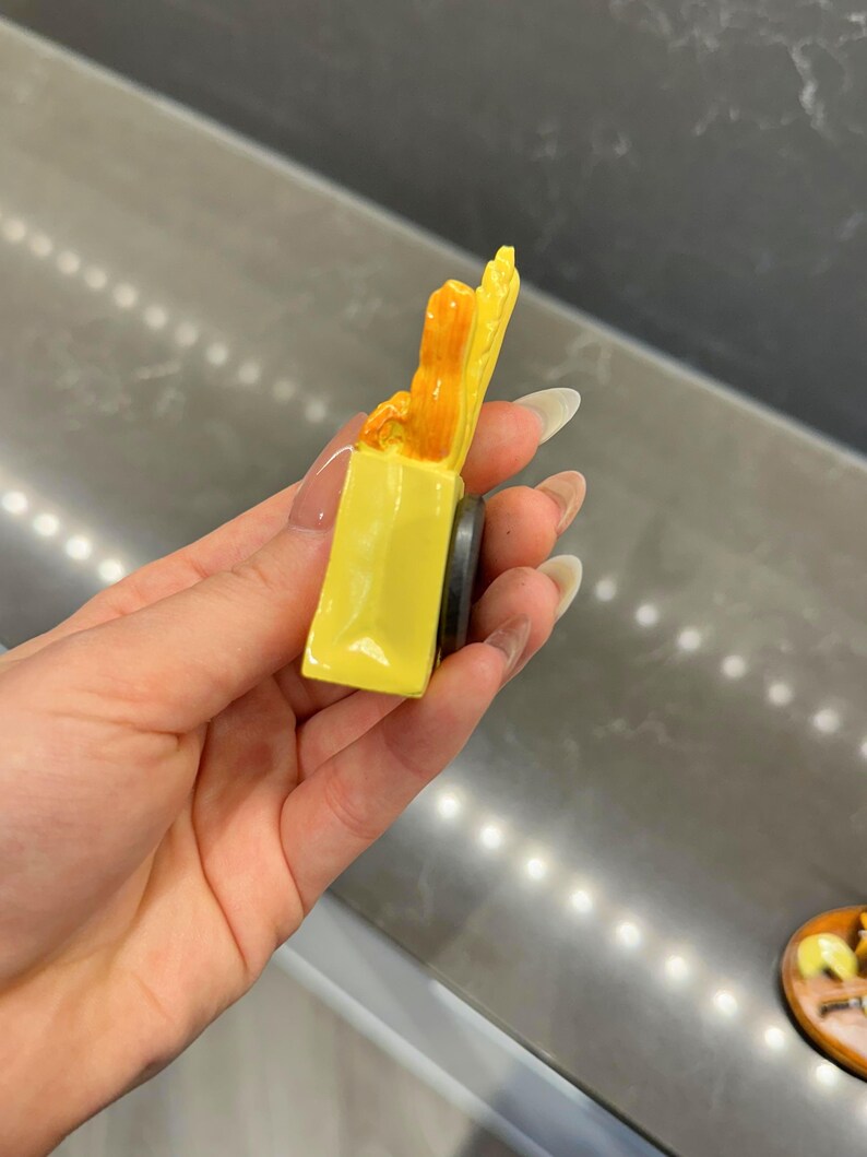 Puede incluir: Un peque&ntilde;o im&aacute;n tridimensional de color amarillo con forma de bolsa de la compra. La bolsa contiene elementos de color naranja que sobresalen por la parte superior. Un disco magn&eacute;tico negro circular est&aacute; fijado a la parte trasera.