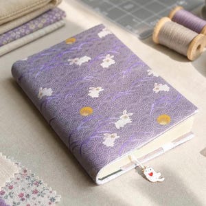 May include: A rectangular notebook with a fabric cover featuring a purple background, white rabbit illustrations, gold circles, and swirling patterns. A white ribbon bookmark with a small white cat charm featuring a red heart is attached.