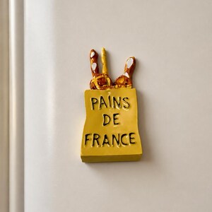 Puede incluir: Un im&aacute;n de nevera rectangular de color amarillo con forma de bolsa de papel y el texto negro PAINS DE FRANCE impreso en el frente. Tres barras de pan tipo baguette de color marr&oacute;n y beige con reflejos blancos sobresalen de la bolsa.