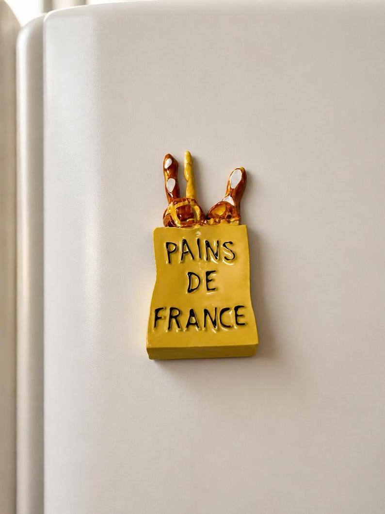 Puede incluir: Un im&aacute;n de nevera rectangular de color amarillo con forma de bolsa de papel y el texto negro PAINS DE FRANCE impreso en el frente. Tres barras de pan tipo baguette de color marr&oacute;n y beige con reflejos blancos sobresalen de la bolsa.