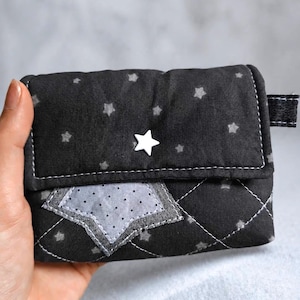 May include: A black quilted fabric wallet featuring a pattern of small light gray stars. The front flap is secured with a white star-shaped snap button and displays a larger, light gray star appliqué with a subtle dark polka dot pattern.