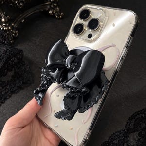 May include: A clear smartphone case featuring a large, glossy black bow attachment with ruffled edges. The case has a light-colored back panel decorated with subtle, thin, curved pink lines and small, scattered, multi-colored confetti-like dots.