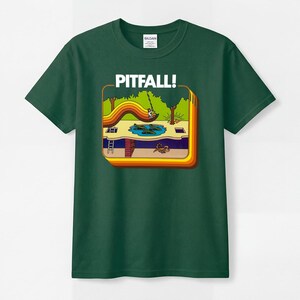 May include: A dark green short-sleeve t-shirt featuring a graphic print of the video game Pitfall! with the text PITFALL! above a scene showing a character swinging over a pool of crocodiles and an underground area with a scorpion.