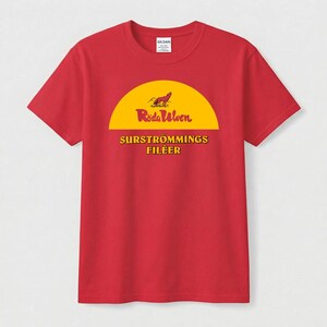 May include: A red short-sleeve t-shirt featuring a yellow semi-circle graphic with a wolf illustration. The text reads "Röda Ulven" and "SURSTRÖMMINGS FILÉER" in a bold, dark font. The shirt has a crew neckline and a Gildan brand label at the neck.