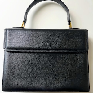 May include: A structured black leather handbag with a textured finish, featuring a single top handle attached with gold-tone hardware. The front flap displays a centered embossed V logo. The bag has a rectangular silhouette with clean, defined edges.