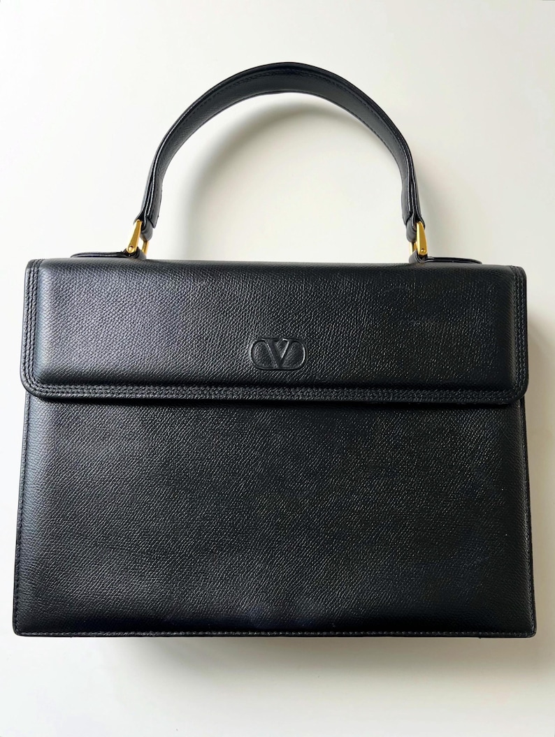 May include: A structured black leather handbag with a textured finish, featuring a single top handle attached with gold-tone hardware. The front flap displays a centered embossed V logo. The bag has a rectangular silhouette with clean, defined edges.