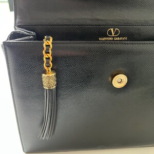 May include: A black leather handbag with a textured finish, featuring a gold-tone metal magnetic snap closure and a decorative gold chain tassel with an engraved cylindrical cap. The interior flap displays the gold text Valentino Garavani.