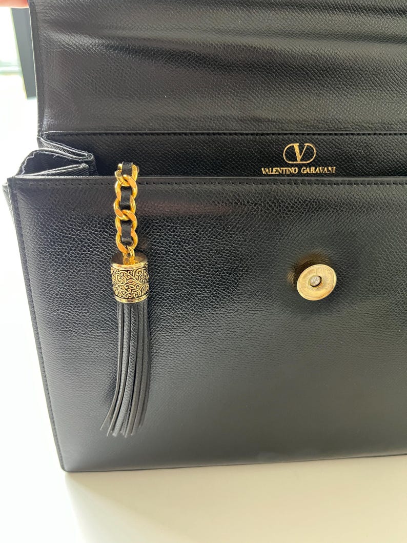 May include: A black leather handbag with a textured finish, featuring a gold-tone metal magnetic snap closure and a decorative gold chain tassel with an engraved cylindrical cap. The interior flap displays the gold text Valentino Garavani.