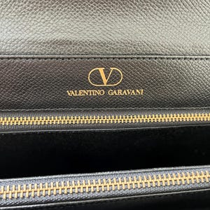 May include: A black textured leather wallet featuring the gold-toned V logo and the text VALENTINO GARAVANI embossed on the interior flap. The wallet includes a gold-toned metal zipper closure with a matching metallic track along the top edge.