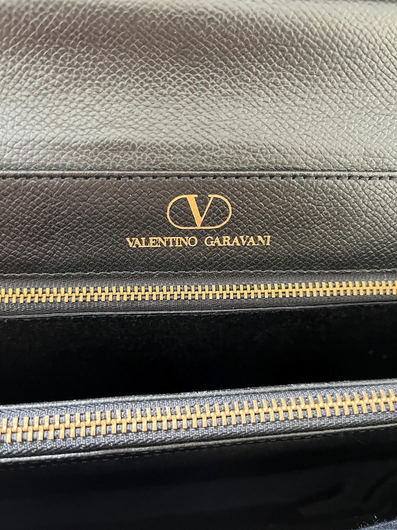 May include: A black textured leather wallet featuring the gold-toned V logo and the text VALENTINO GARAVANI embossed on the interior flap. The wallet includes a gold-toned metal zipper closure with a matching metallic track along the top edge.