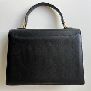 May include: A structured black handbag with a textured surface, featuring a single top handle attached with gold-tone metal hardware. The bag has a rectangular silhouette with a flap closure and clean, minimalist lines throughout the design.