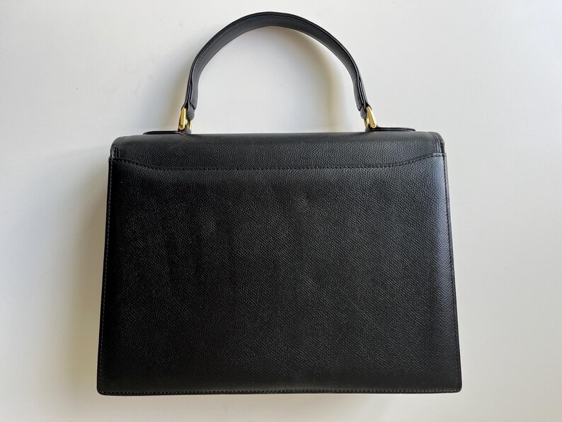May include: A structured black handbag with a textured surface, featuring a single top handle attached with gold-tone metal hardware. The bag has a rectangular silhouette with a flap closure and clean, minimalist lines throughout the design.