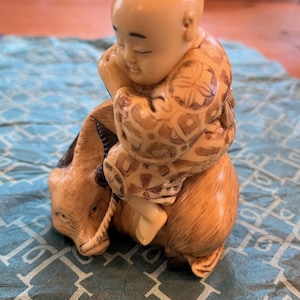 May include: A detailed figurine of a sleeping child riding a goat. The child is wearing a patterned garment. The goat has a rope around its neck. The figurine is primarily beige and brown, with intricate details.