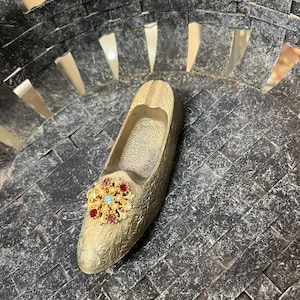 Puede incluir: Cenicero dorado en forma de zapato con un adorno ornamental tipo joya. El cenicero tiene una superficie texturizada y está colocado dentro de una cesta tejida gris oscuro.