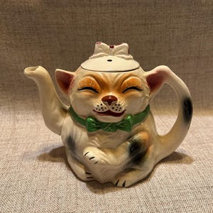 May include: A ceramic teapot shaped like a cat with a cream-colored body, black and brown patches, and a green bow tie. The cat has a smiling expression with closed eyes, a brown nose, and a white bow on the lid. A curved handle and spout extend from the sides.