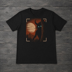 May include: A black short-sleeved t-shirt featuring a graphic print of a dark, horned creature with glowing cyan eyes peering from behind a tree. The scene includes a large moon, mountains, and pine trees, framed by camera viewfinder markings.