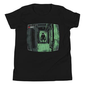 May include: A black short-sleeved t-shirt featuring a graphic print of a security camera view down a hallway. The green-tinted image shows a dark silhouette with glowing eyes and a wide grin, with the text 23:48:12 10/26 and a red REC indicator.