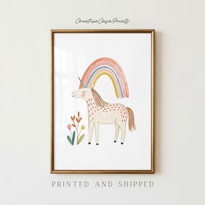 Puede incluir: Una lámina de arte enmarcada con una ilustración en acuarela de un unicornio de color crema con crin y cola multicolor, de pie bajo un arcoíris pastel. Cerca de los pies del unicornio se observan pequeñas flores estilizadas.