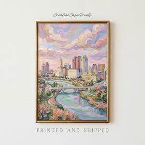 Könnte beinhalten: Ein Ölgemälde in einem goldenen Rahmen zeigt eine Skyline mit einem Fluss und einer Brücke im Vordergrund. Der Himmel ist mit dicken, strukturierten Pinselstrichen in Rosa-, Lavendel- und Pfirsichtönen gefüllt, die sich im Wasser und an Gebäuden spiegeln.