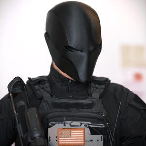 May include: A matte black full-face helmet with a smooth, contoured design and narrow eye slits. The helmet is worn with a black tactical vest featuring a rectangular patch displaying an American flag in orange and black tones.