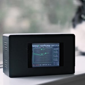 May include: A rectangular black electronic device with a digital screen displaying a radar interface. The screen shows the text HUMAN/NON-HUMAN ENTITY, telemetry data, and three heart icons with distance measurements in feet, including 5.2ft, 1.5ft, and 2.0ft.