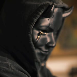 May include: A dark gray hooded sweatshirt worn with a black mask featuring two prominent horns on the forehead and a metallic gold tear-like detail extending from the right eye socket. The mask has a smooth finish and covers the face completely.