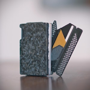 May include: A compact wallet featuring a dark gray marbled carbon fiber exterior plate. The wallet is partially open, revealing internal card slots, a black textured frame with a perforated pattern, and a single gold-colored card insert.