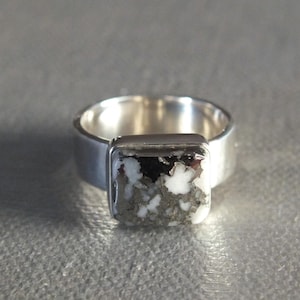 May include: A silver ring with a rectangular stone. The stone features a mix of white, black, and gray colors. The ring band is a simple, wide design.
