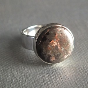 May include: A silver ring with a large, round, dark stone. The stone has a mottled appearance with shades of brown, gray, and copper. The ring band is thick and polished, reflecting the light.