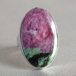 Può includere: Anello ovale in argento con una gemma lucida. La pietra presenta una miscela di colori rosa, verde e nero. Il design dell'anello è semplice, evidenziando la bellezza naturale della pietra.
