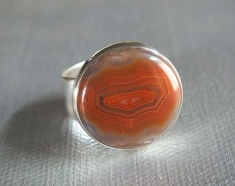 May include: A silver ring featuring a large, round agate stone. The stone displays concentric bands of orange, red, and white, creating a striking pattern. The ring's band is a simple, polished silver.
