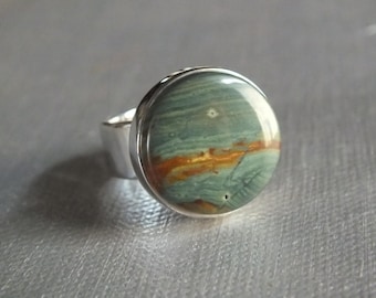 Op de afbeelding: Zilveren ring met een grote, ronde steen. De gepolijste steen toont een mix van groene, bruine en oranje tinten, waardoor een natuurlijk patroon ontstaat. De ring heeft een eenvoudige, brede band.