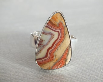 May include: A silver ring with a large, teardrop-shaped stone. The stone is a mix of brown, orange, and white with a swirling pattern.