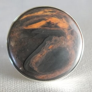 Può includere: Pomello decorativo rotondo per mobili con base in metallo color argento. La superficie del pomello presenta un motivo a vortice di tonalità marrone scuro e arancione, creando un effetto marmorizzato. Il design ricorda la pietra naturale.