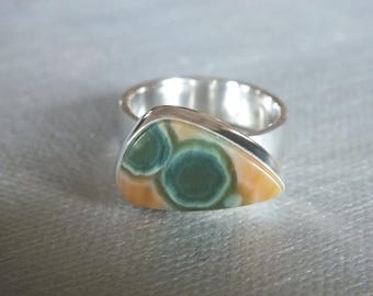 Op de afbeelding: Een zilveren ring met een grote, driehoekige edelsteen. De steen heeft een patroon van groene en bruine kleuren. De ringband is van eenvoudig, gepolijst zilver.