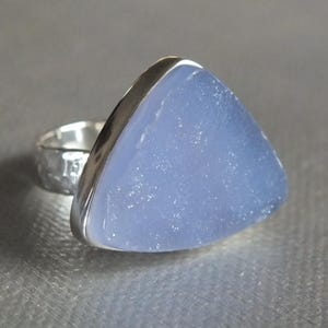 May include: A silver ring featuring a large, triangular, pale blue gemstone. The gemstone has a slightly textured surface and is set in a polished silver bezel. The ring band has a hammered texture.