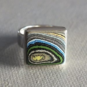 May include: A silver ring with a square, multi-coloured stone. The stone features layers of black, white, blue, green, and yellow, creating a unique pattern. The ring has a simple, polished band.
