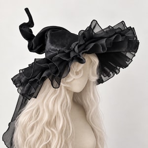May include: A black pointed hat featuring a tall, slightly curved crown made of velvet-like fabric. The wide brim is adorned with multiple layers of sheer, ruffled organza trim that extends into a long, flowing veil-like piece at the back.