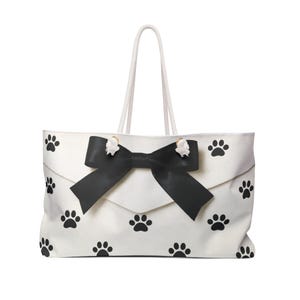 Puede incluir: Un bolso tote blanco con un patrón repetitivo de huellas de patas negras. El bolso tiene un gran lazo negro centrado en la solapa frontal y dos asas de cuerda blanca sujetas con herrajes metálicos, ofreciendo un diseño estructurado.