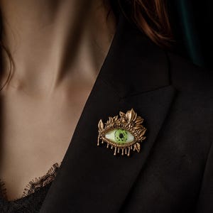 May include: A bronze-toned brooch shaped like an eye with a bright green iris and a black pupil. The eye is surrounded by ornate, leaf-like metal detailing and features small, dangling metal drops along the bottom edge of the iris.