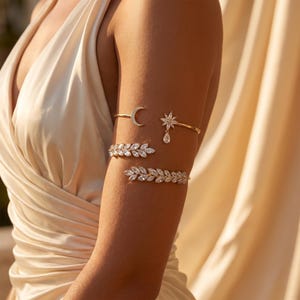 Op de afbeelding: Een set van drie goudkleurige armbanden voor de bovenarm met heldere kristallen. De bovenste armband heeft een halve maan en een ster met een druppelvormig accent. De middelste en onderste armbanden hebben een bladvormig patroon van kristallen.