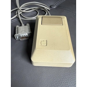 May include: A vintage beige Apple computer mouse with a single rectangular button and a long gray cable ending in a D-sub connector. The mouse features a molded Apple logo on the top surface and has a textured, rectangular plastic casing.