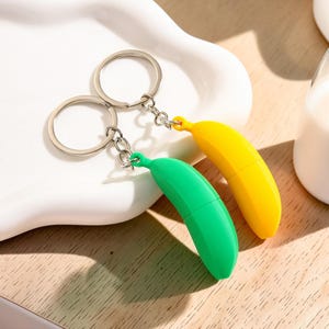 May include: Two keychains featuring banana-shaped charms, one in bright yellow and one in vibrant green. Each charm is attached to a silver-toned metal split ring by a short chain. The banana charms have a smooth, solid-coloured plastic finish.