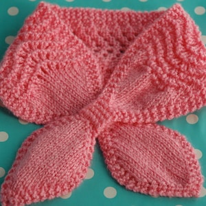 Puede incluir: Una bufanda de punto rosa con un diseño de lazo. La bufanda está hecha con un punto texturizado y tiene un aspecto delicado y femenino.