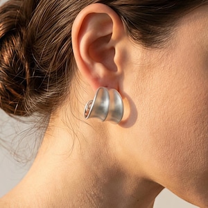 May include: A silver-toned earring with a curved, semi-circular shape and a smooth, matte finish. The earring features a double-layered design that creates a hollow, arching structure, securely positioned on the earlobe with a visible metal backing.