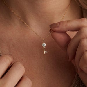 May include: A gold chain necklace featuring a small key-shaped pendant. The pendant is designed with a round, clear, faceted stone set in the circular head of the key and a tiny, square, clear stone embedded in the lower shaft of the key.
