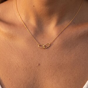 Puede incluir: Un collar de tono dorado con una cadena fina y un colgante central compuesto por dos pequeños eslabones ovalados entrelazados. El collar descansa sobre la clavícula, mostrando un diseño sencillo y refinado con un acabado metálico pulido.
