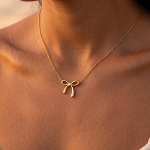 May include: A gold-toned necklace featuring a delicate bow-shaped pendant suspended from a fine, thin chain. The pendant is centered on the chain, which rests against the collarbone, showcasing a smooth, polished metallic finish throughout the piece.