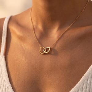 May include: A gold-toned necklace featuring two interlocking heart-shaped pendants suspended from a delicate chain. The metal has a smooth, polished finish, and the two hearts are linked together to form a single central focal point on the chain.